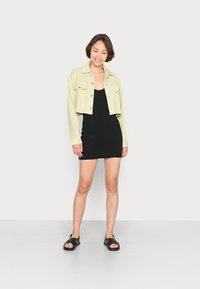 Casaco de cortiça verde claro, cortado, sobre um vestido preto de malha, combinado com sandálias pretas. O fundo é branco liso. Look casual.