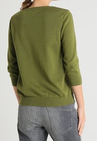 Olivgrüner Pullover mit Rundhalsausschnitt, Dreiviertelärmeln und geripptem Saum, kombiniert mit grauen Jeans mit Gesäßtaschen.