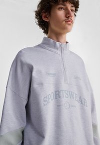 Ljusgrå zip-up sweatshirt i mjukt tyg, med hög krage, logodetaljer och "SPORTSWEAR" tryckt i blått. Färgblockerade ärmar.