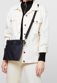 Borsa a tracolla blu navy realizzata in tessuto morbido, con una tasca frontale e una tracolla regolabile, appoggiata su una giacca di colore chiaro.