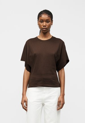 Fonott hajú nő karika fülbevalóval, laza, sötétbarna rövid ujjú felsőben és fehér nadrágban, egy egyszínű világos háttér előtt állva.