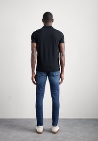 Man som står med ryggen mot, klädd i en svart kortärmad pikétröja, blå jeans och vita sneakers på ett grått golv.