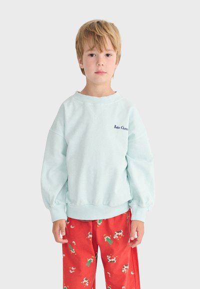 Enfant blond portant un sweat-shirt bleu clair avec le texte « Bobo Chose » et un pantalon rouge orné d'illustrations colorées de vaches et de fermiers, debout immobile.