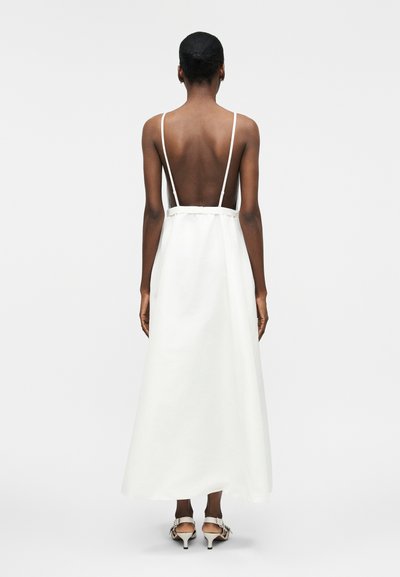 Γυναίκα που φαίνεται από πίσω, φοράει ένα λευκό φόρεμα χωρίς πλάτη με λεπτές τιράντες και λευκά σανδάλια με τακούνι, στέκεται μπροστά σε απλό ανοιχτόχρωμο φόντο.