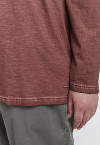 Una camicia rossa a maniche corte, texture con taglio dritto, dotata di un piccolo logo a fondo e polsini cuciti. Indossata con pantaloni grigi.