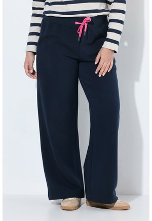 Persona che indossa pantaloni larghi blu navy con coulisse rosa, camicia a righe a maniche lunghe e scarpe beige, in piedi contro un muro bianco.