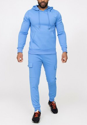 Homme portant un sweat à capuche bleu clair et un pantalon cargo de jogging assorti, avec des baskets noires aux lacets orange, debout devant un fond blanc.