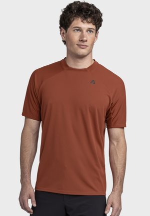 Mann mit kurzen lockigen Haaren, der ein rostfarbenes kurzärmeliges Sportshirt und schwarze Hosen trägt, schaut leicht nach links.