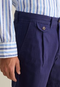 Helles blau-weiß gestreiftes Hemd mit Knopfmanschetten, kombiniert mit navyblauen Hosen mit gefälteltem Design und Knopfverschluss.