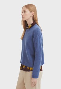 Blå stickad tröja med rundad hals och sänkta axlar, lagd över en blommig skjorta med krage. Utsmyckningen kompletteras med beige byxor.