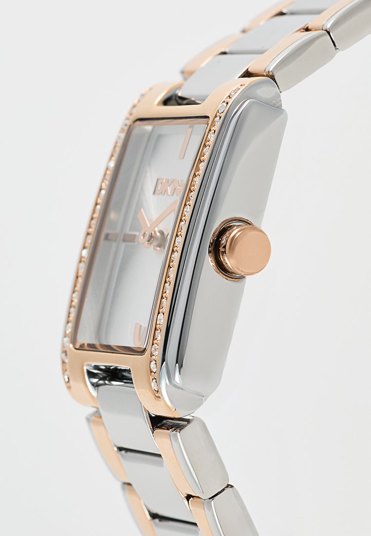 Prostokątny srebrno-różowo-złoty zegarek na rękę z pierścieniem wysadzanym kryształami, różowo-złotą koronką i metalową bransoletą w ogniwach.