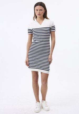 Kvinde iført hvid og marineblå kortærmet kjole med krave og hvide sneakers mod hvid baggrund.