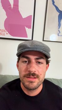 Szürke lapos sapka, fekete pulóver, látható bajusz. Világos bőrszín. A háttérben két művészeti plakát látható, rózsaszín és kék mintákkal.