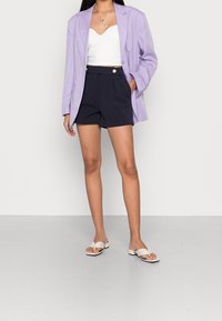 Fialový oversize blazer, bílý zkrácený top, námořnické šortky s vysokým pasem a zlatými knoflíky, a nahé sandály na klínku. Jednoduchý, čistý design.