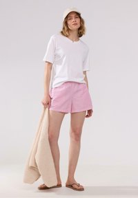Vit bomull t-shirt, rosa och vita randiga shorts, håller en beige stickad filt, bär en beige keps och bruna flip-flops.