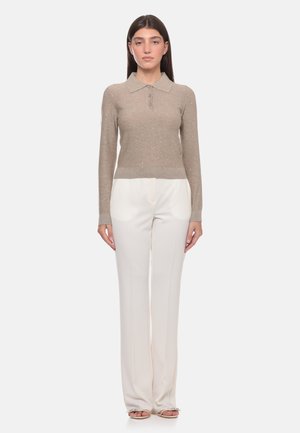 Donna in piedi che indossa un maglione beige a maniche lunghe con colletto e pantaloni bianchi a gamba larga con sandali aperti su sfondo bianco.