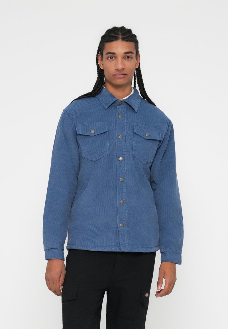 Blå cordurojakkesæt med knaplukning foran, to brystlommer og en klassisk krave. Stoffet har en struktureret, ribbet overflade.