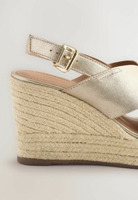 Sandalette à talon compensé avec une tige en cuir doré métallique et une plateforme en jute beige texturé. Bride de cheville réglable avec une boucle dorée.