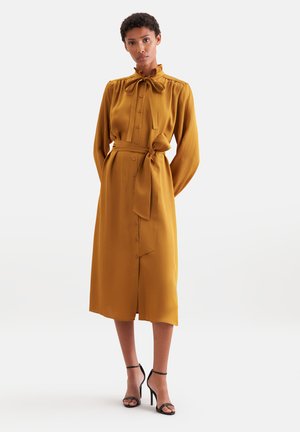 Femme portant une robe midi jaune moutarde à manches longues avec des boutons, une ceinture à la taille et des sandales noires à talons hauts, debout sur un fond blanc.