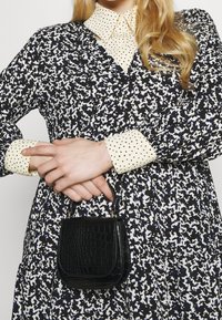 Black crocodile-textured mini handbag with a top handle. Worn with a patterned black-and-white dress featuring a collared cream shirt.