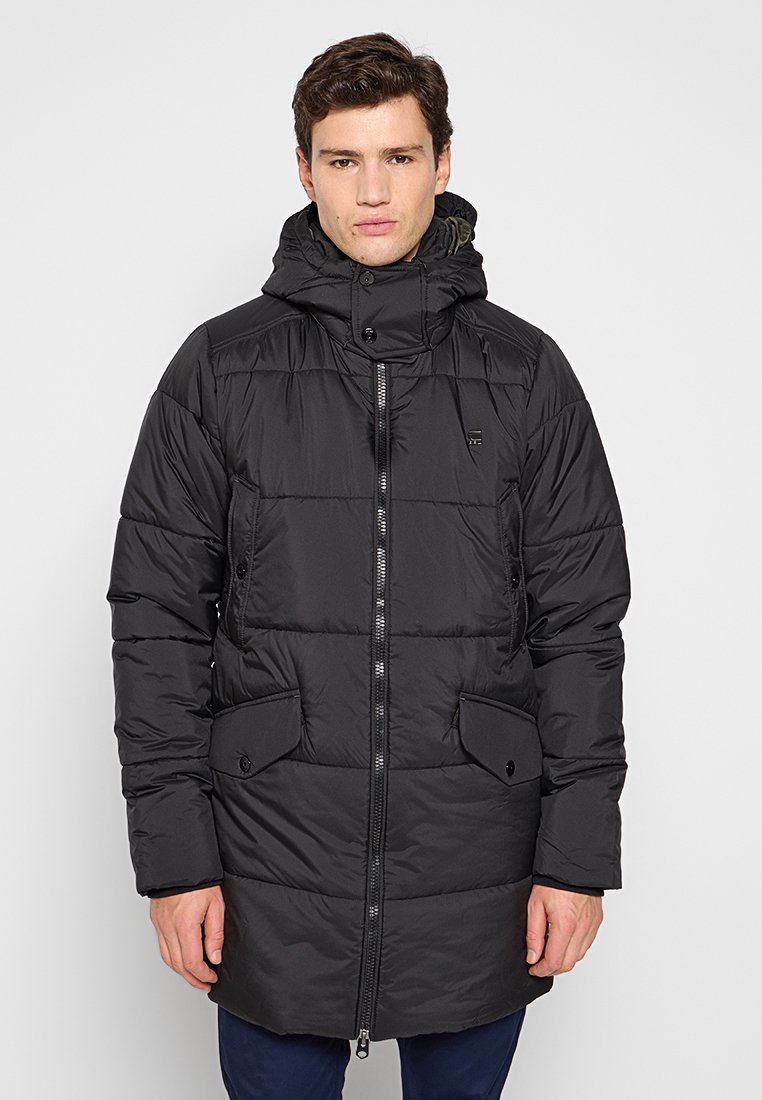 Hombre vestido con un abrigo de invierno negro largo y acolchado, con capucha, cremallera frontal, bolsillos con solapa abotonados, y de pie frente a un fondo liso.