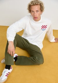 Valge pikkade varrukatega shirt punase ja sinise ruudulise logoga, koos oliiviroheliste pükstega ja retrostiilis valgete tossudega punaste aktsentidega.