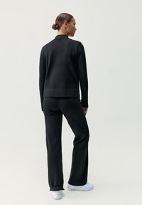 Svart, texturerad jacka med rak passform, ihop med matchande byxor. Modellen bär vita skor, ses bakifrån mot en ljus bakgrund.