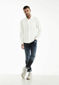 Bílá košile s dlouhým rukávem a přední kapsou, v kombinaci s tmavými džíny a bílými teniskami. Hladký materiál a volný střih.