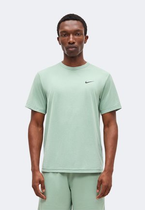 Mann stående, iført lys grønn Nike t-skjorte og matchende shorts, nøytral uttrykk, ensfarget lys bakgrunn.
