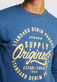 Blå bomulls sweatshirt med rund halsringning. Har stor gul text med texten "SUPPLY Originals" och annan branding i en cirkulär layout.