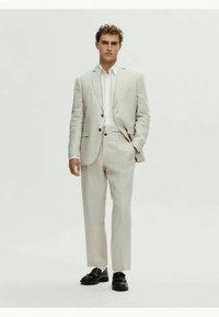 Ανοιχτό γκρίζο λινό κοστούμι με σακάκι ενός κουμπιού, λευκό πουκάμισο και μαύρα παπούτσια. Οι παντελόνες είναι ίσιες με κομμένη εφαρμογή.