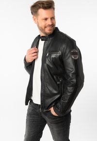 Zwart leren motorjack met een hoge kraag, voorrits en een ritsvak op de borst. Bevat een ronde patch op de linkerschouder.