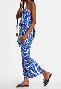 Blå mönstrad byxdress med volangliv, vida ben och svarta sandaler med text. Materialet verkar lätt och luftigt.