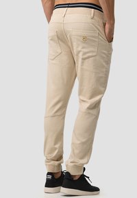 Pantalons de jogging beige avec des poignets élastiques, deux poches arrière et un logo. Associés à des baskets noires avec un dessus texturé et une semelle blanche.