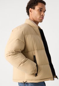 Jachetă puffer, bej, cu interior negru, având un guler înalt, buzunare cu fermoar și manșete elastice pentru căldură și confort.