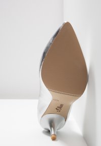 Chaussure à talon haut argentée à bout pointu vue par la semelle, avec semelle beige texturée et talon fin de 5 cm.