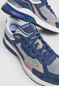 Las zapatillas deportivas en azul marino y beige cuentan con materiales de ante y malla, una lengüeta texturizada y una suela de goma blanca con una marca en el costado.