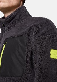 Szara polarowa kurtka z wysokim kołnierzem posiada czarną kieszeń na piersi z materiału oraz neonowożółty zamek. Zawiera naszywkę z logo na rękawie.