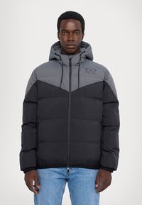 Giacca imbottita in nero e grigio con cappuccio, trapuntatura verticale, zip frontale e logo EA7 sulla parte superiore del petto destro.
