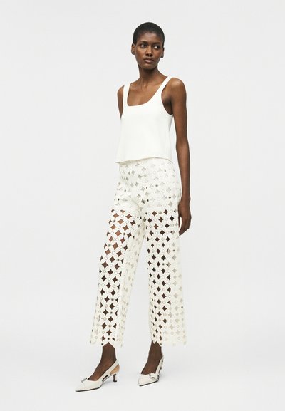 Femme portant un haut blanc sans manches et un pantalon large en crochet avec des motifs en losange, assorti à des chaussures blanches à talons bas.