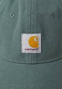 Grønn stofflue med et frontpatch som har en oransje og hvit logo. Strukturert overflate med synlig søm og avrundet brem.