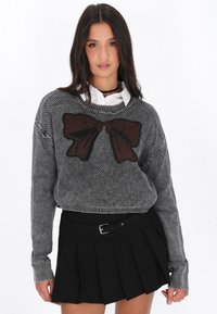 Grauer Strickpullover mit einem braunen Schleifenmotiv, gerippte Bündchen und Saum, kombiniert mit einem schwarzen plissierten Rock mit einem Gurt-Detail.