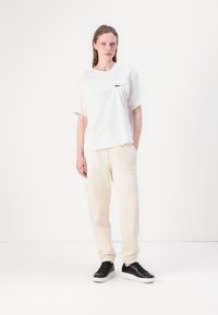 Vit bomullströja med en grön logotyp på bröstet, kombinerad med krämfärgade träningsoverallsbyxor och svarta sneakers. Enkla, avslappnade linjer.