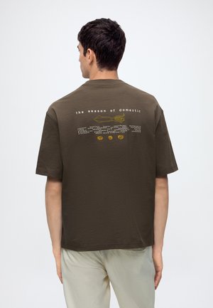 Homme portant un t-shirt vert olive foncé avec des textes et des graphiques blancs et jaunes dans le dos, associé à un pantalon beige clair.