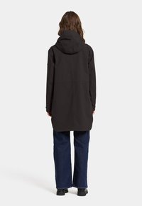 Svart vattentät jacka med huva, medellång, med en slät yta och två sidofickor. Matchad med lösa blå jeans och svarta skor.