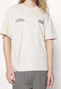 Beżowa bawełniana koszulka z czarnym napisem: "PALACZE UMIERAJĄ MŁODO" po lewej stronie oraz "PEDUS" na piersi. Krótkie rękawy i luźny krój.