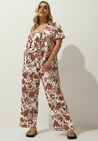 Jumpsuit a stampa floreale bianco con foglie e fiori marroni, con bottoni frontali, maniche corte e pantaloni a gamba larga.
