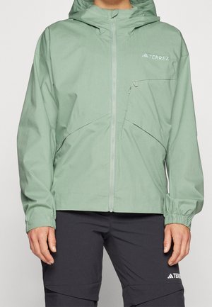Persona que lleva una chaqueta Terrex con capucha de color verde claro con cremallera frontal y bolsillo con cremallera en el pecho, combinada con pantalones negros.