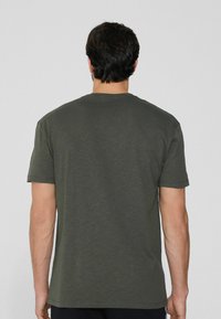 Oliwkowa koszulka z krótkim rękawem, okrągłym dekoltem i miękkim, teksturowanym materiałem. Tył jest gładki i prosty, z luźnym krojem.