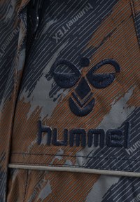 Graues und orangefarbenes gemustertes Gewebe mit einem gestickten Logo in symmetrischem Design. Das Material hat eine glatte Textur.